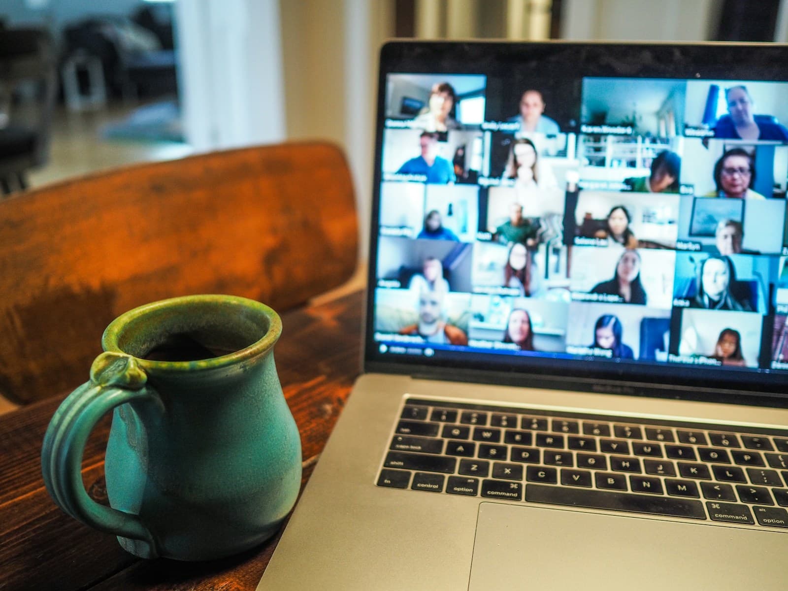 A laptop on a wooden table showing a video call with a grid of participants, next to a green ceramic mug