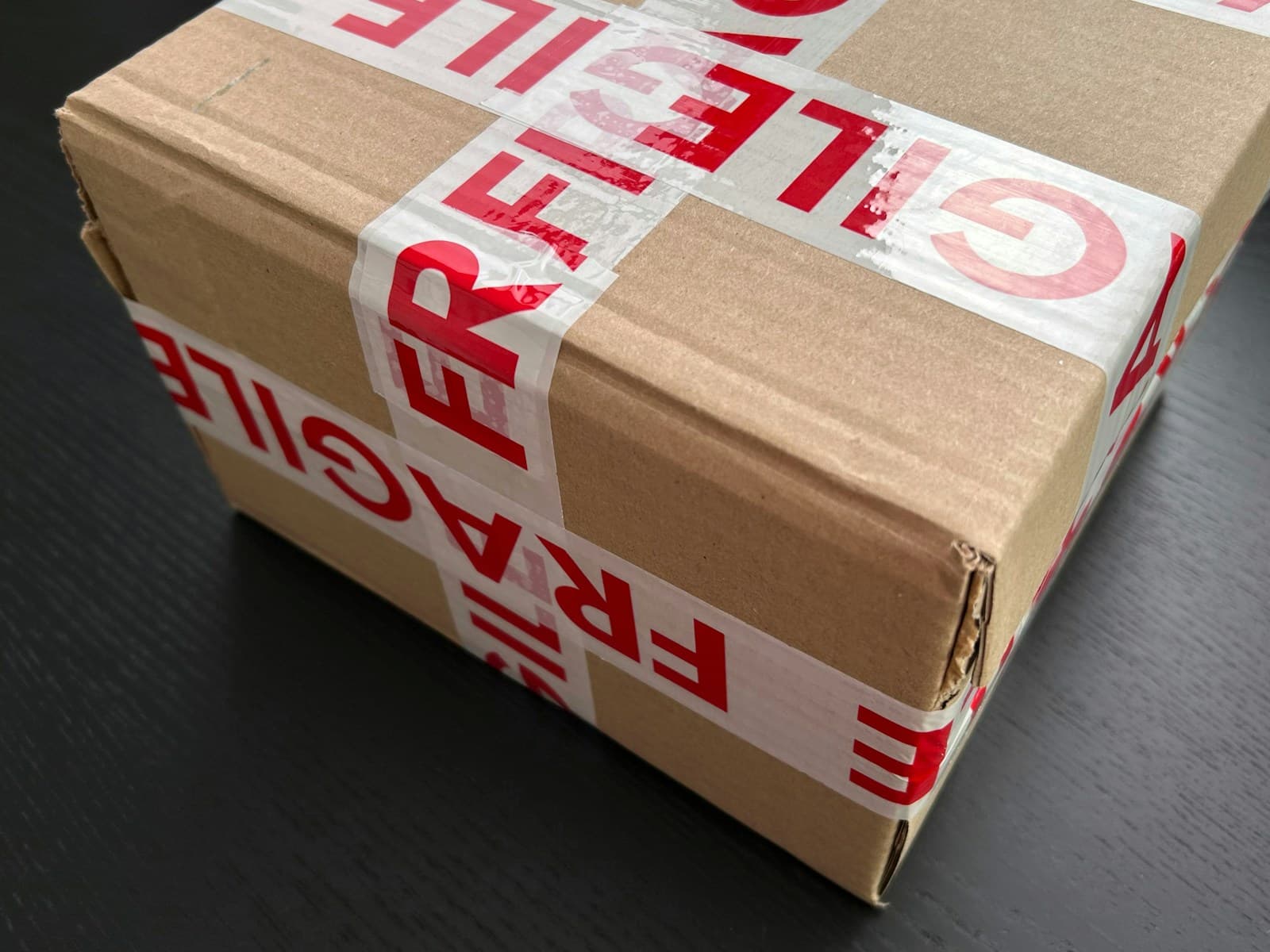 A brown cardboard parcel wrapped in red and white FRAGILE tape, with one corner of the box crushed and torn