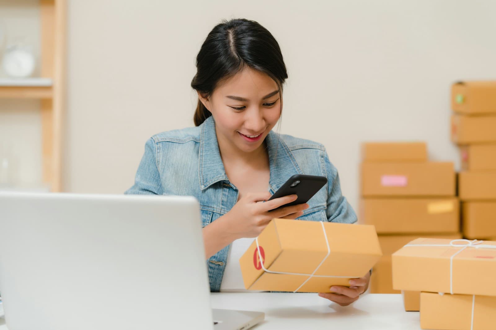 Smiling woman in a denim jacket checking her phone while holding a wrapped parcel, with a laptop and stacked shipping boxes beside her