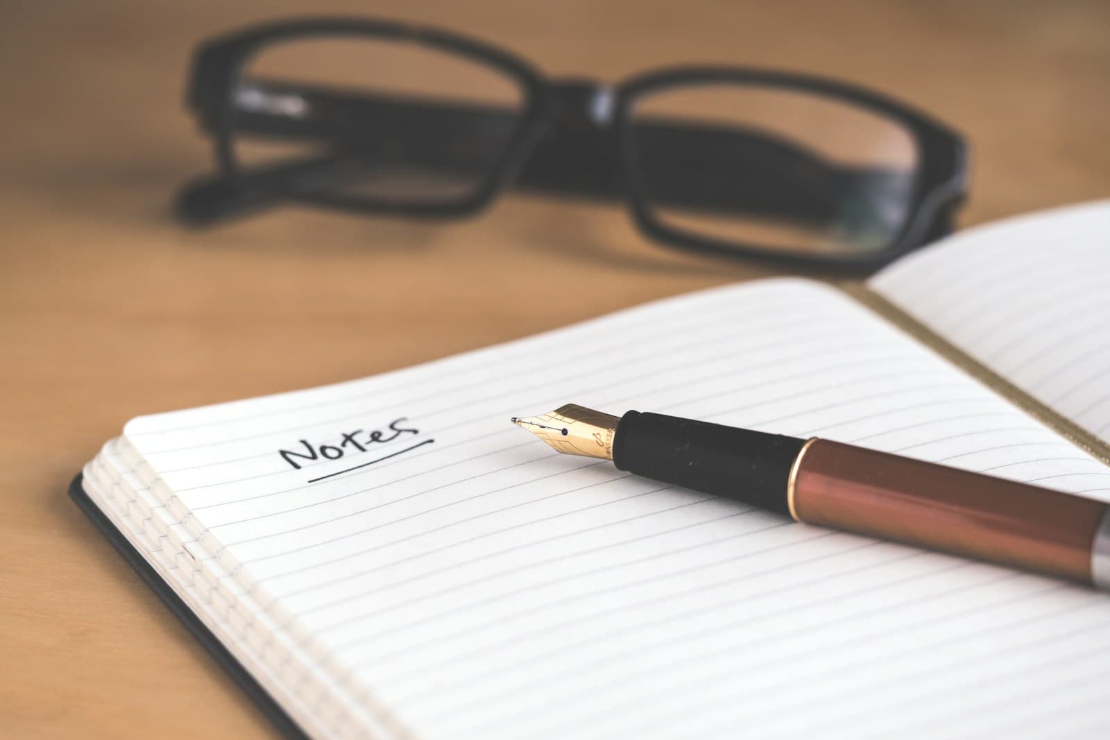 An open lined notebook with the word Notes handwritten at the top, a fountain pen resting on the page and reading glasses behind it